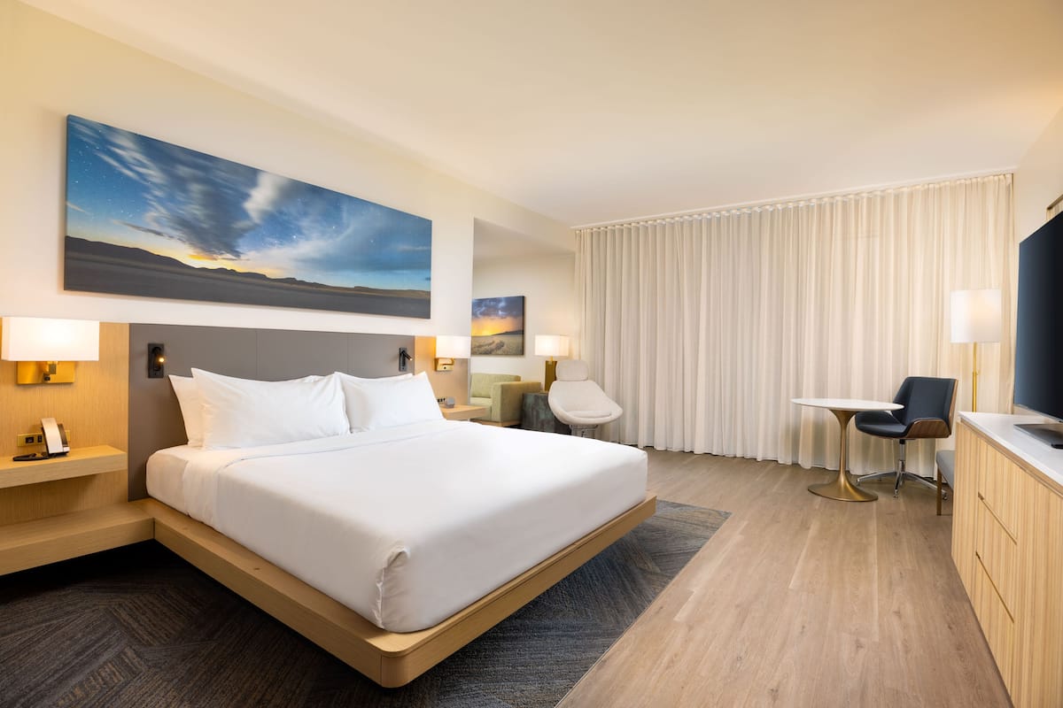 Ein Hotelzimmer mit einem großen Bett, Nachttischen, einem Schreibtisch mit Stuhl und Blick zum Himmel durch das Fenster.