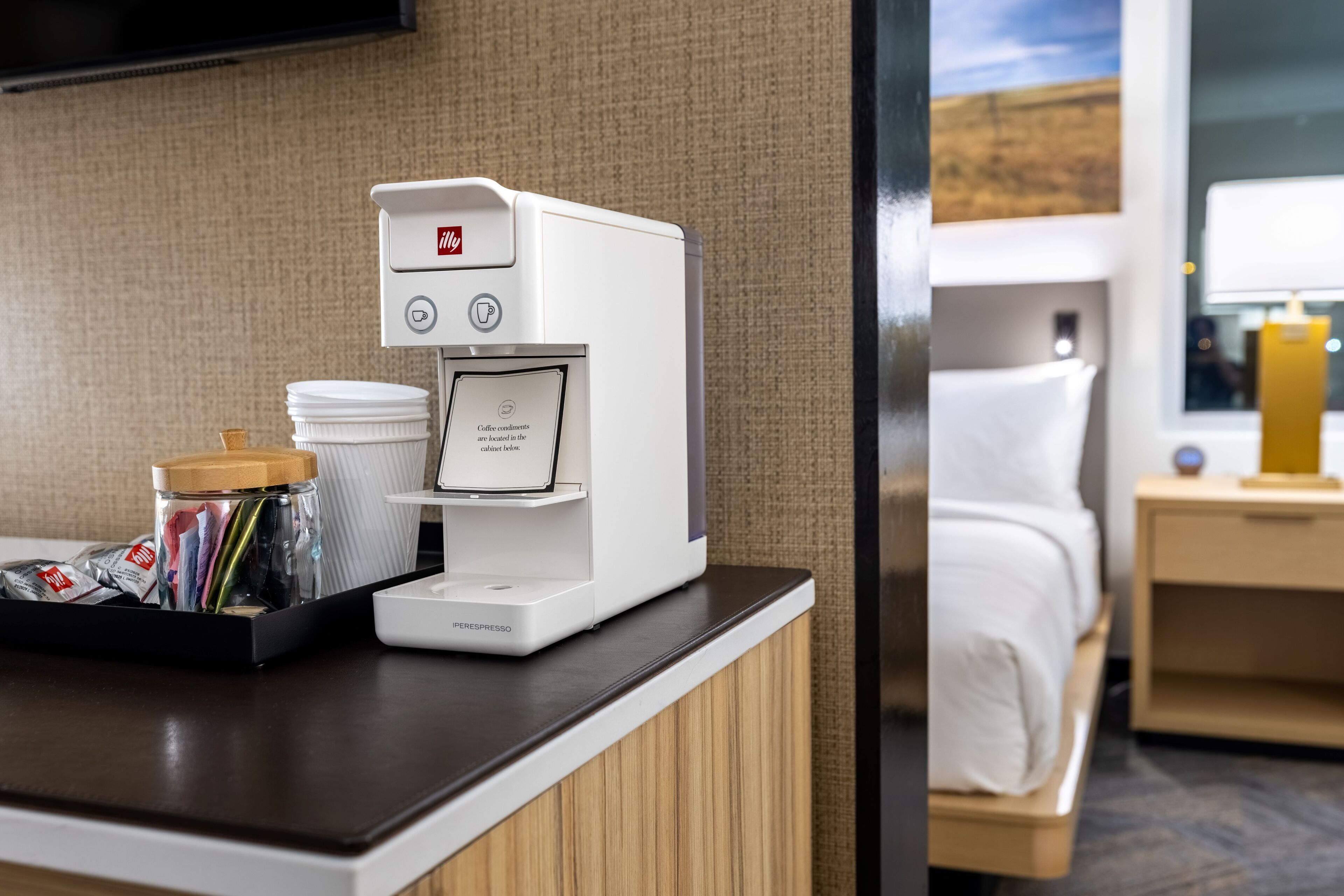 A hotel room with a coffee machine, a tray with cups and a jar of coffee pods, and a bed with a nightstand.