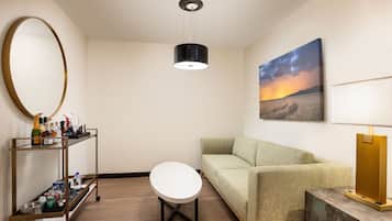 Una habitación con un espejo redondo, una mesa blanca, un sofá, una estantería con botellas, un cuadro de un atardecer y una lámpara.