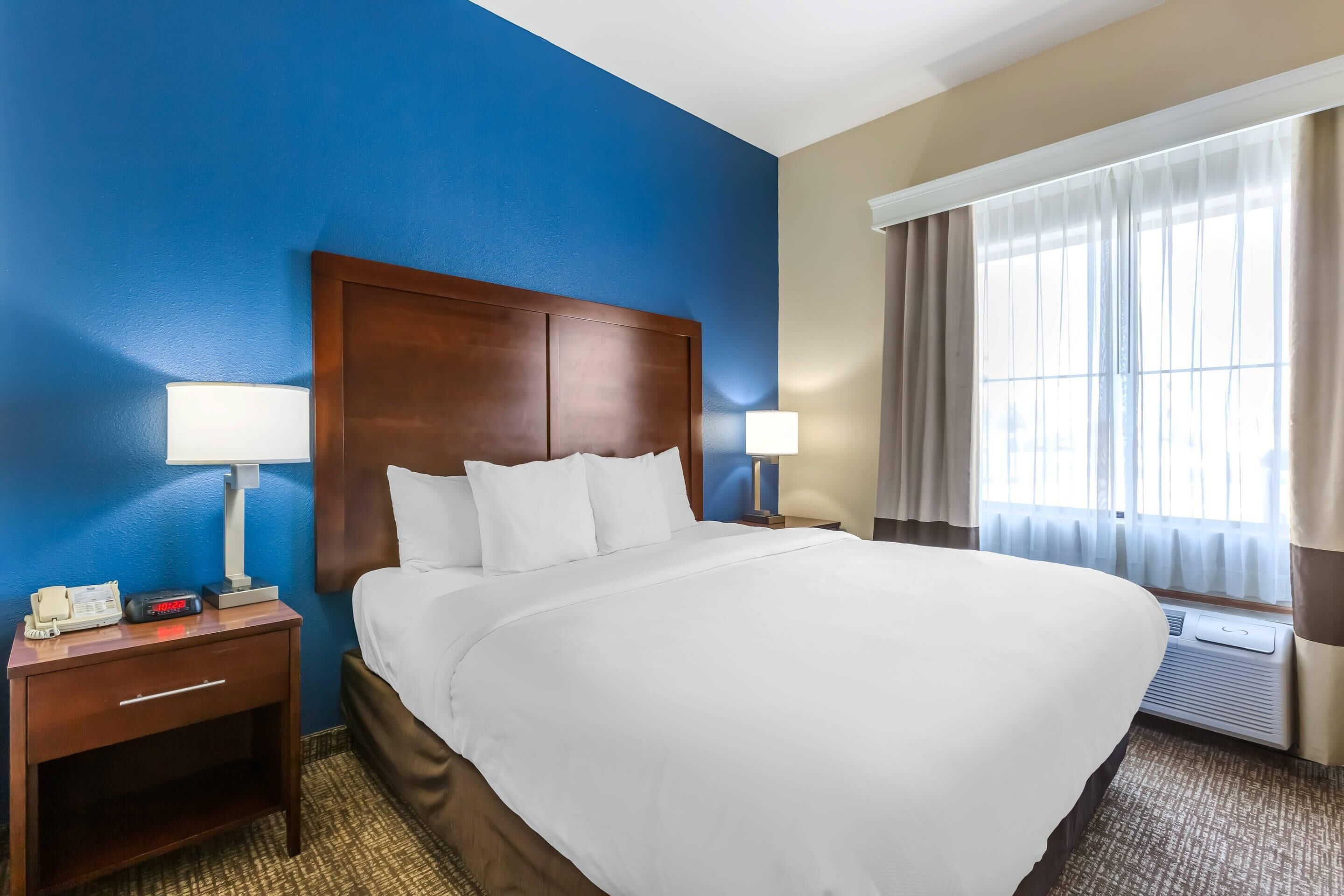 Une chambre d’hôtel avec un grand lit, deux tables de chevet avec des lampes, une petite table avec un téléphone et une fenêtre avec des rideaux.