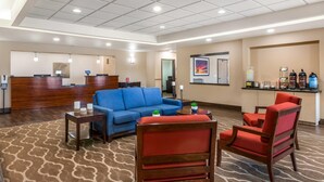 Eine Hotelhalle mit einer Rezeption, einem blauen Sofa, roten Sesseln, einer Kaffeestation und einem gerahmten Bild an der Wand.