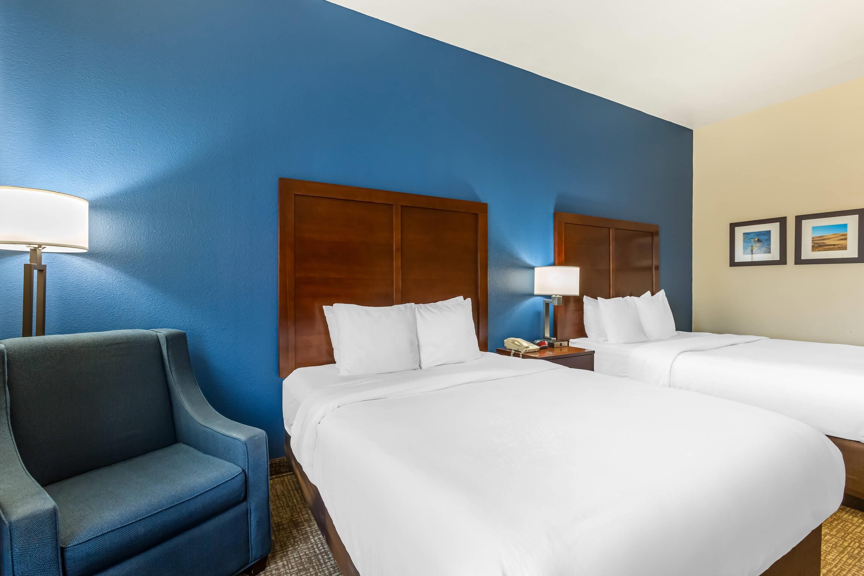 A hotel room with two beds, a desk, a chair, and a lamp.