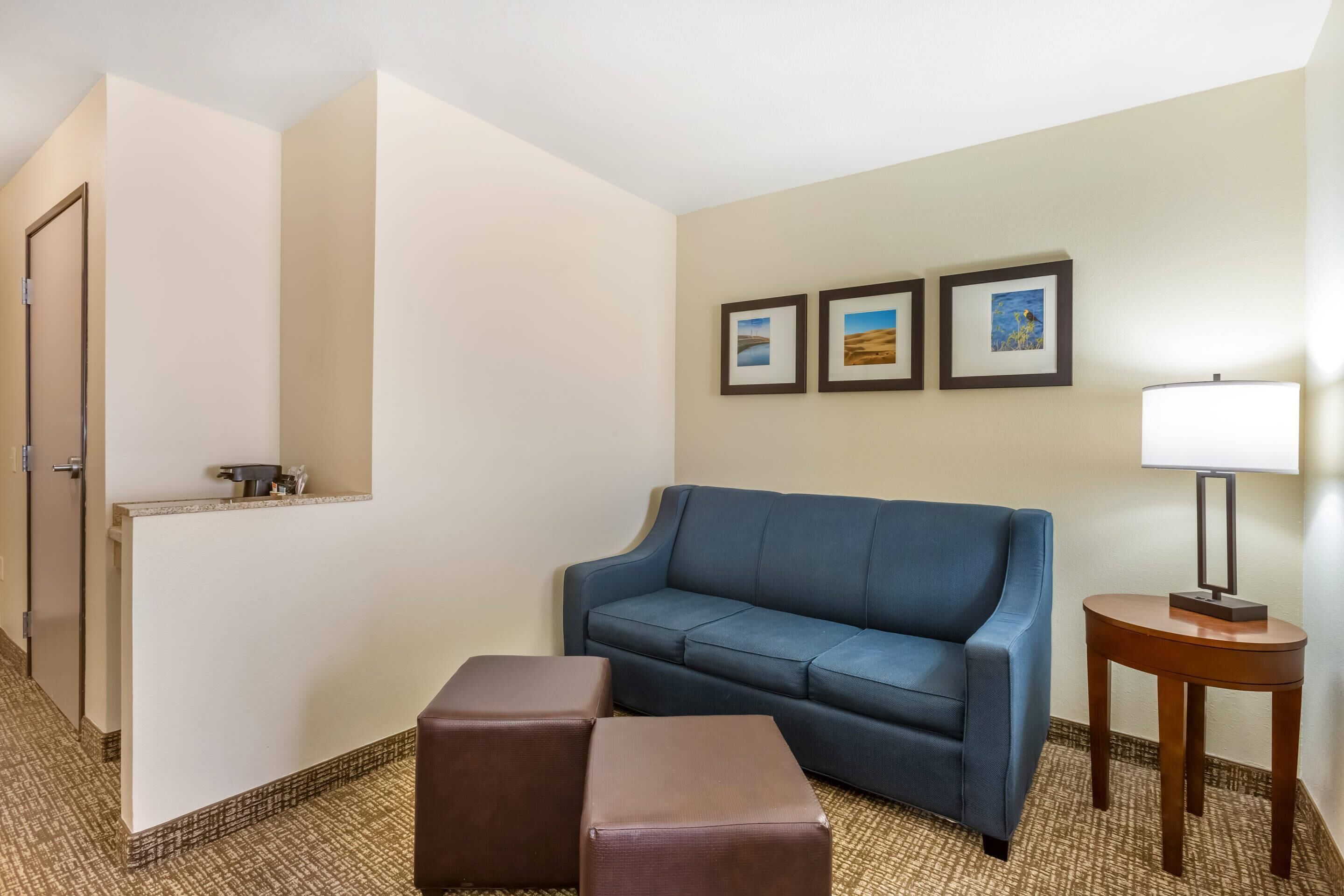 A hotel room with a blue sofa, two brown ottomans, a small table with a lamp, and three framed pictures on the wall.