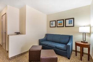A hotel room with a blue sofa, two brown ottomans, a small table with a lamp, and three framed pictures on the wall.