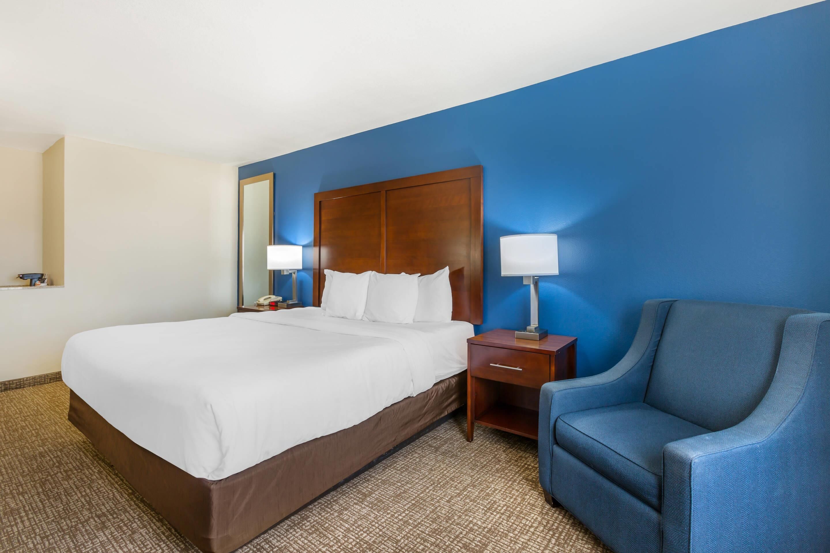 A hotel room with a large bed, a wooden headboard, a blue armchair, a nightstand, and a mirror.