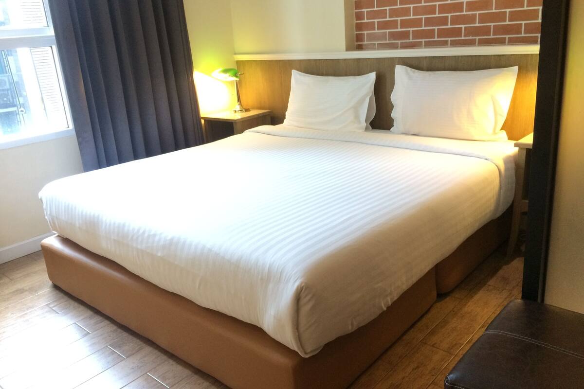 A large bed with a wooden frame and a white mattress, a nightstand with a lamp, and a brick wall behind the bed.