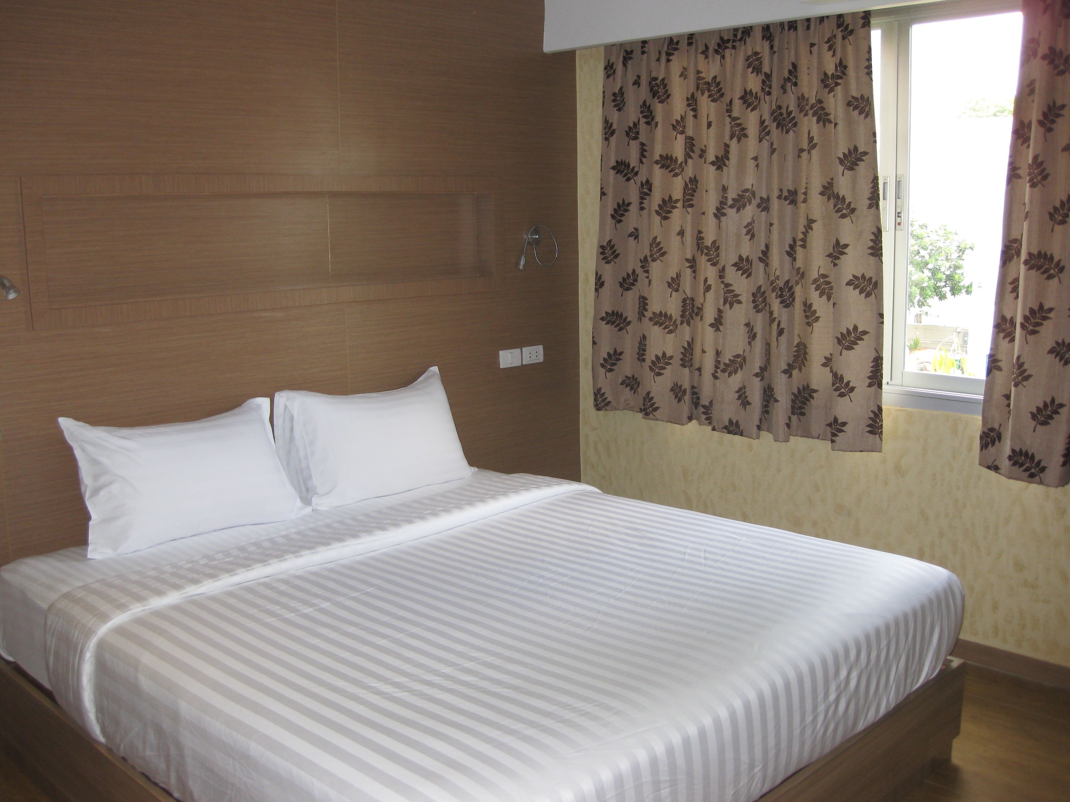 A neatly made bed with white linens in a room with wooden paneling and a window with floral curtains.