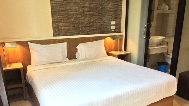 Habitación de hotel con una cama grande, cabecero de madera, dos mesitas de noche con lámparas, pared con textura de piedra y vistas al baño, que incluye un lavabo e inodoro.