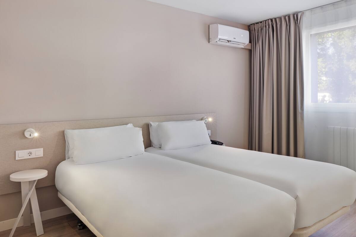 Habitación de hotel con dos camas, una mesita de noche, un equipo de aire acondicionado y una ventana con cortinas.