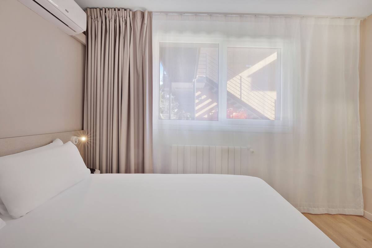 Una habitación de hotel con una cama grande, una ventana con cortinas traslúcidas, un radiador y un equipo de aire acondicionado.