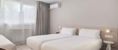 Habitación de hotel con dos camas, una mesita de noche pequeña, una silla, una ventana con cortinas, un equipo de aire acondicionado y una lámpara fijada a la pared.