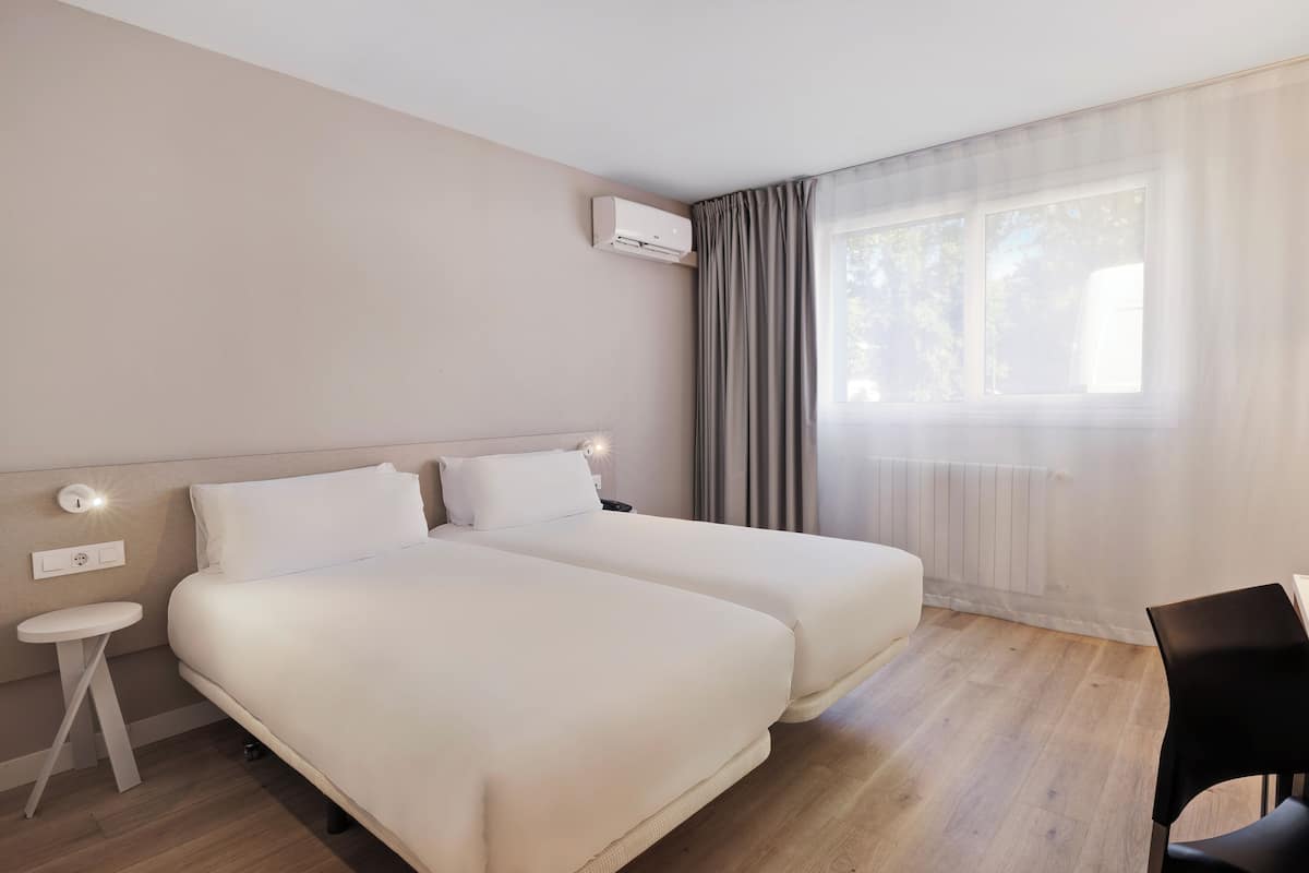Habitación de hotel con dos camas, una mesita, una silla y un ventanal con cortinas.