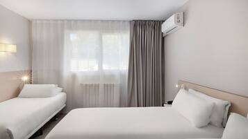 A hotel room with two single beds, a large window with sheer curtains, a wall-mounted air conditioner, and a bedside table with a lamp.