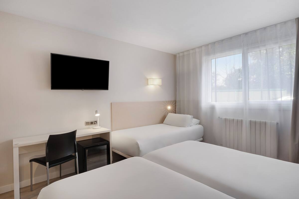 Habitación de hotel con televisión de pantalla plana en la pared, un escritorio con una silla, dos camas y una ventana con cortinas de gasa.
