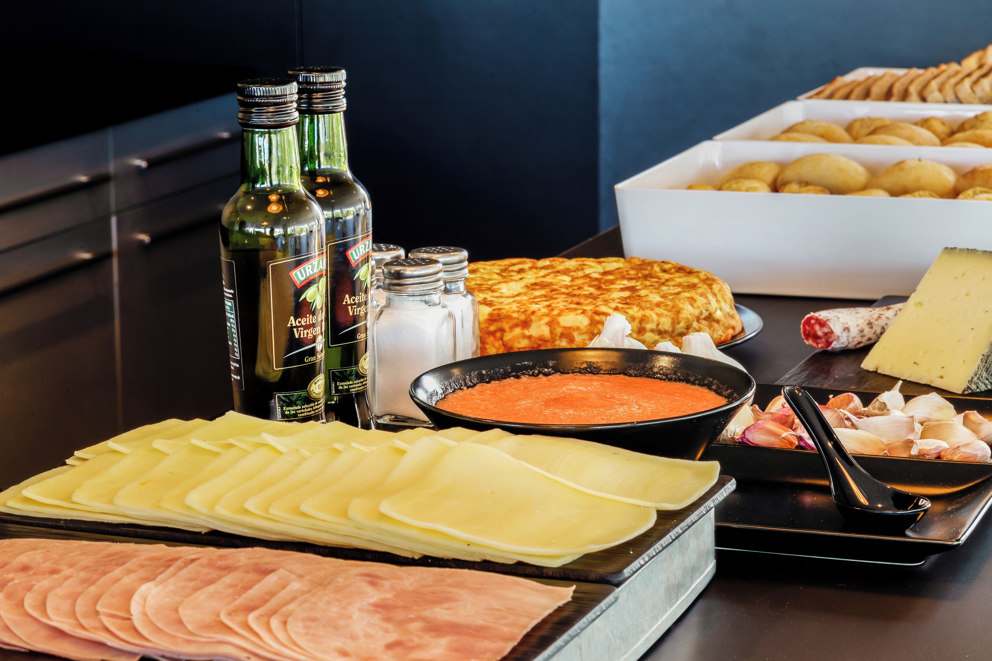 A buffet table with various food items including cheese, ham, and a dish with a red sauce. There are also bottles of wine and salt and pepper shakers.