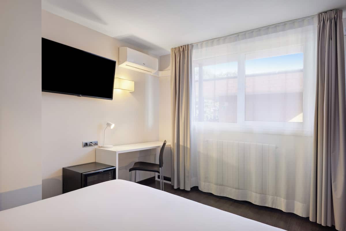 Habitación de hotel con cama, escritorio, silla, televisión de pantalla plana y ventana con cortinas.