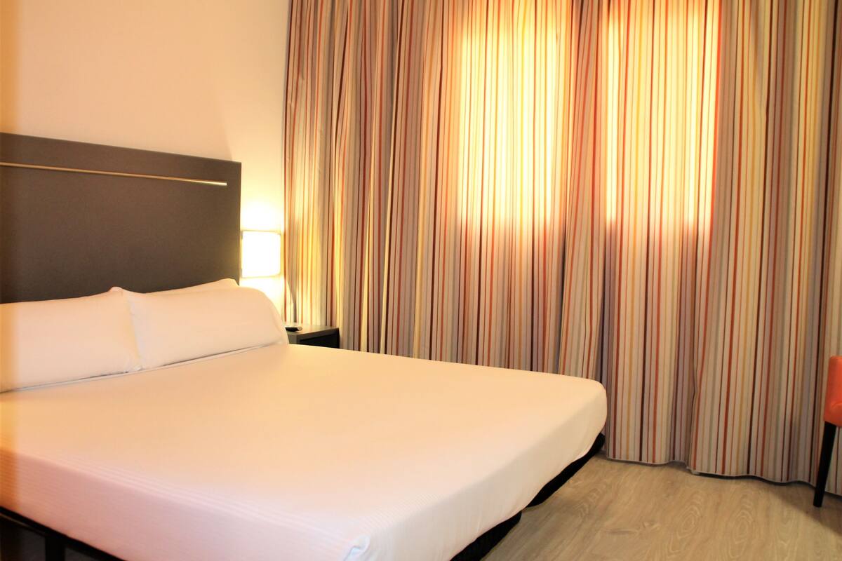 A hotel room with a large bed, a nightstand, a lamp, and a window with curtains.