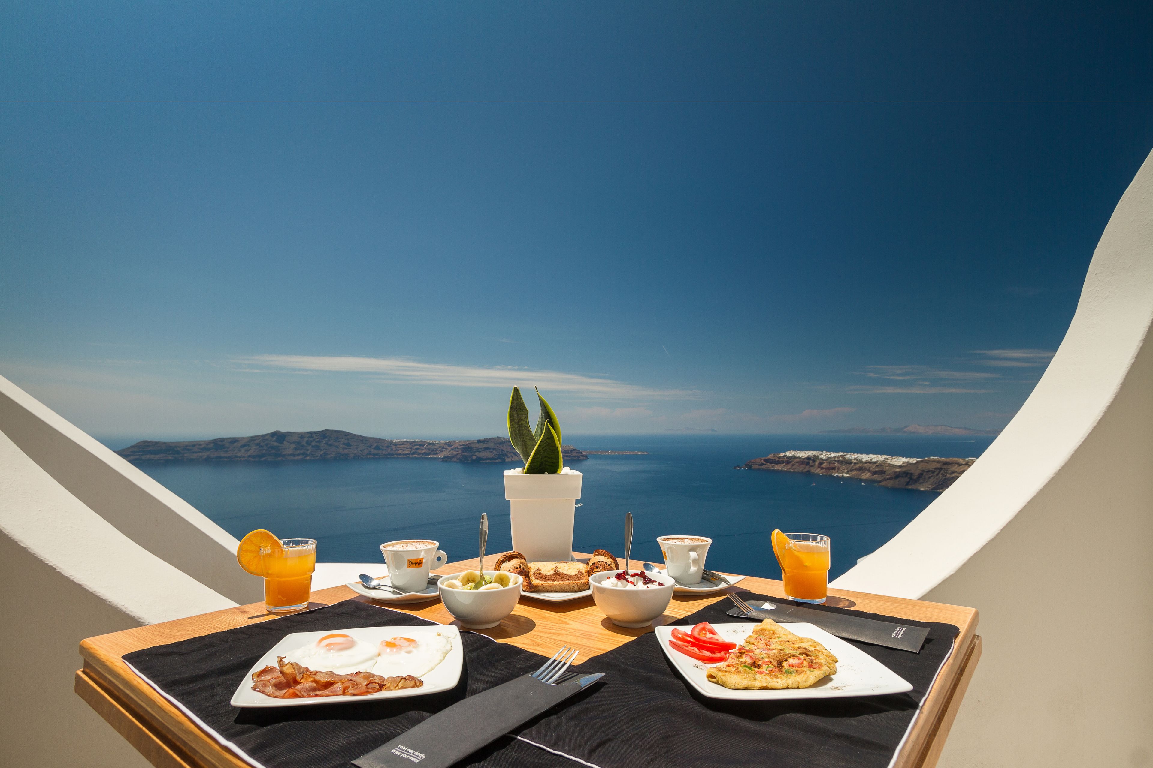 A breakfast table with various dishes, a potted plant, and a view of the sea.