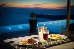A table with a meal, a candle, and a glass of red drink, overlooking a pool and a sunset view. - Galaxy Suites & Villas (Santorini)