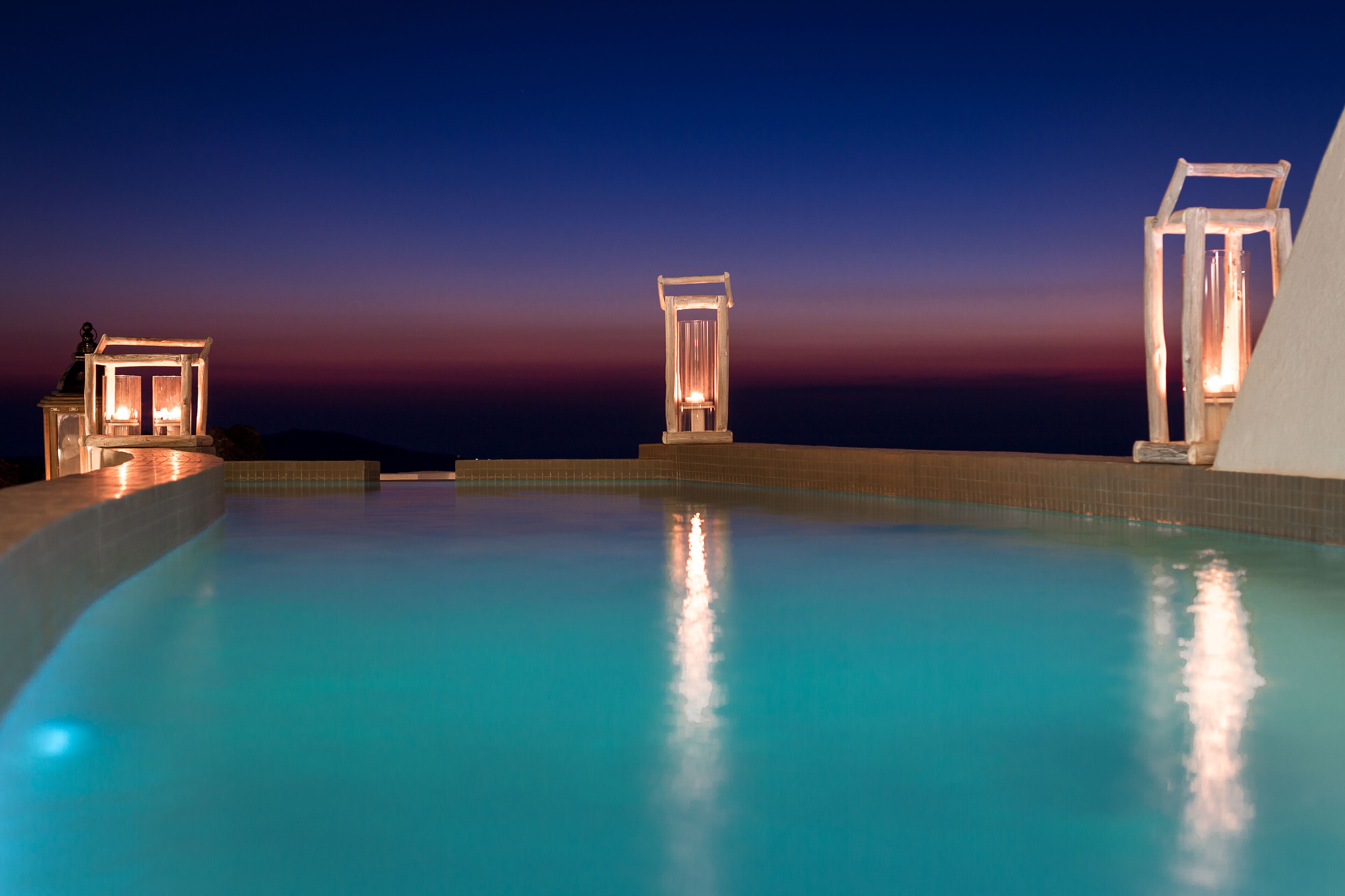Piscina com água cristalina, rodeada por um deck revestido e iluminada por luzes.