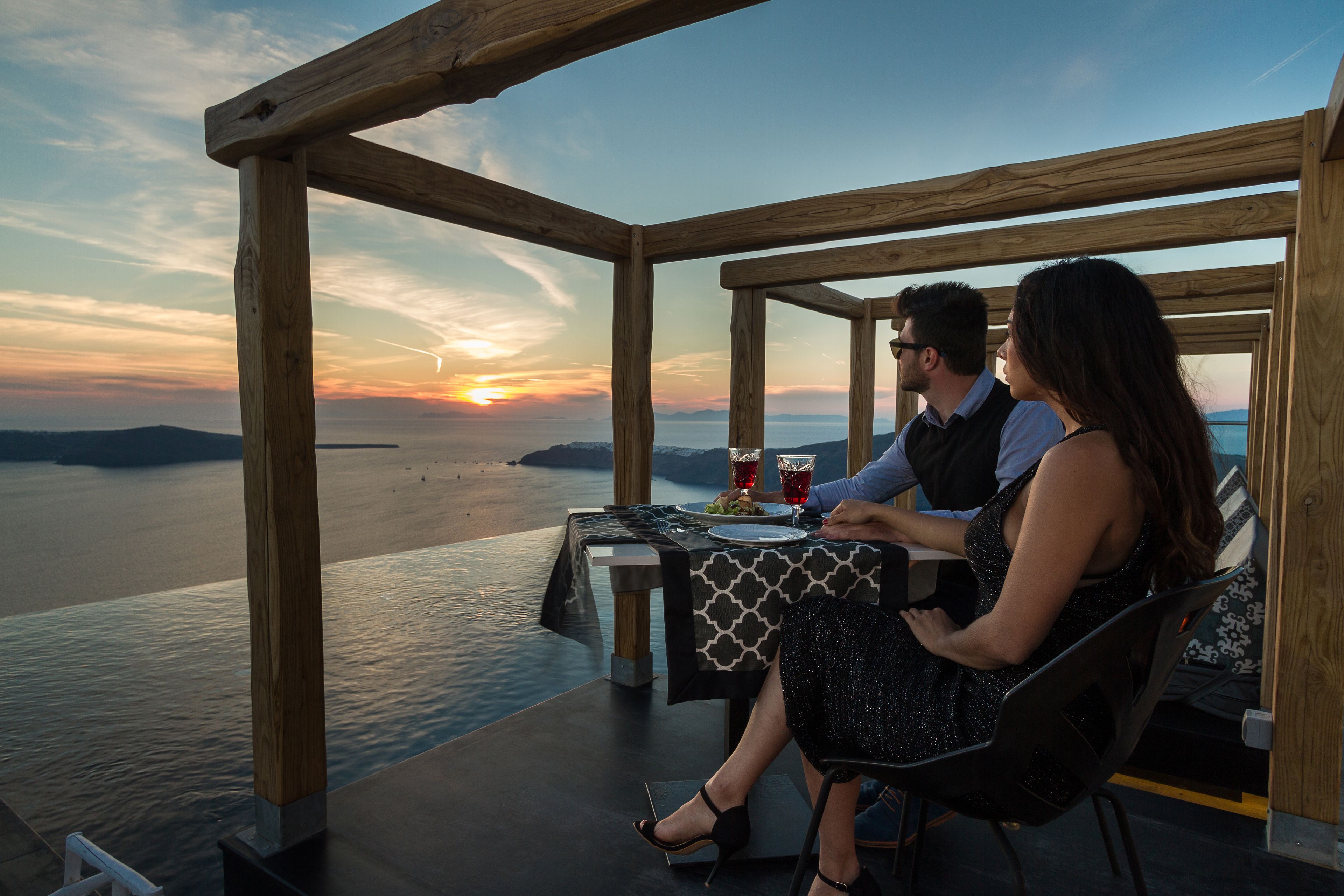 Un couple est assis à une table en plein air, profitant de la vue sur le coucher de soleil qui se reflète sur l’eau.