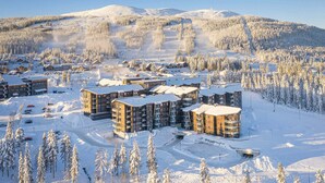 Estância de esqui coberta de neve com vários edifícios, incluindo um hotel de destaque e um teleférico visível ao fundo.