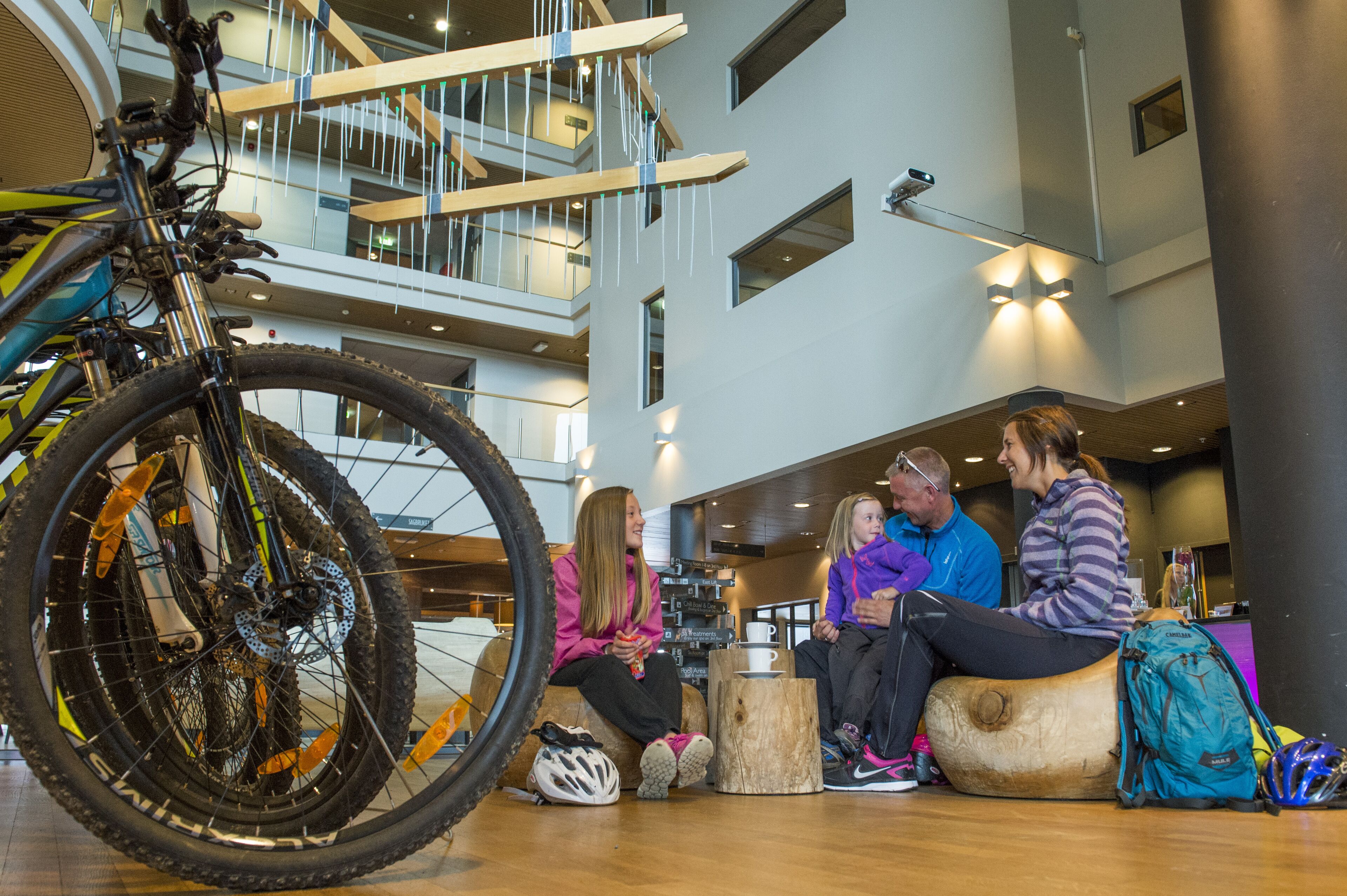 A group of people sitting on wooden stools in a modern indoor space with a large bicycle wheel in the foreground.