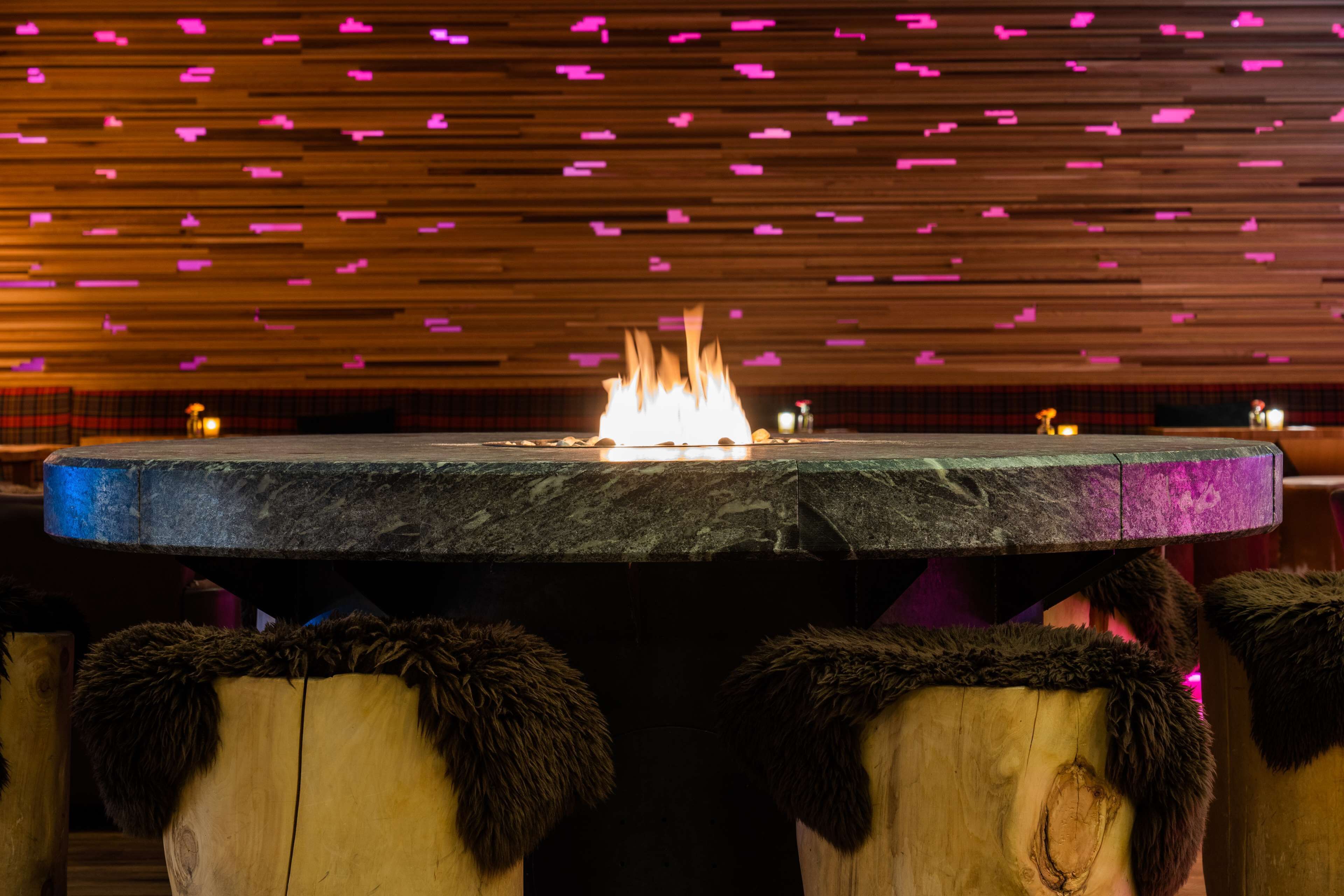 A round table with a central fire, surrounded by wooden stools with fur trim.