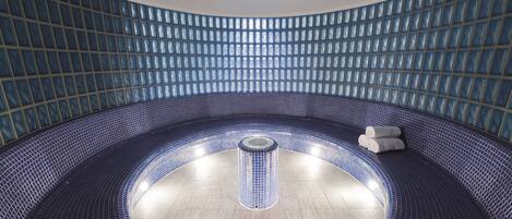 A modern indoor swimming pool with a curved design, featuring a central hot tub area and a tiled wall with a grid pattern.
