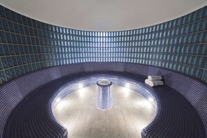 A modern indoor swimming pool with a curved design, featuring a central hot tub area and a tiled wall with a grid pattern.