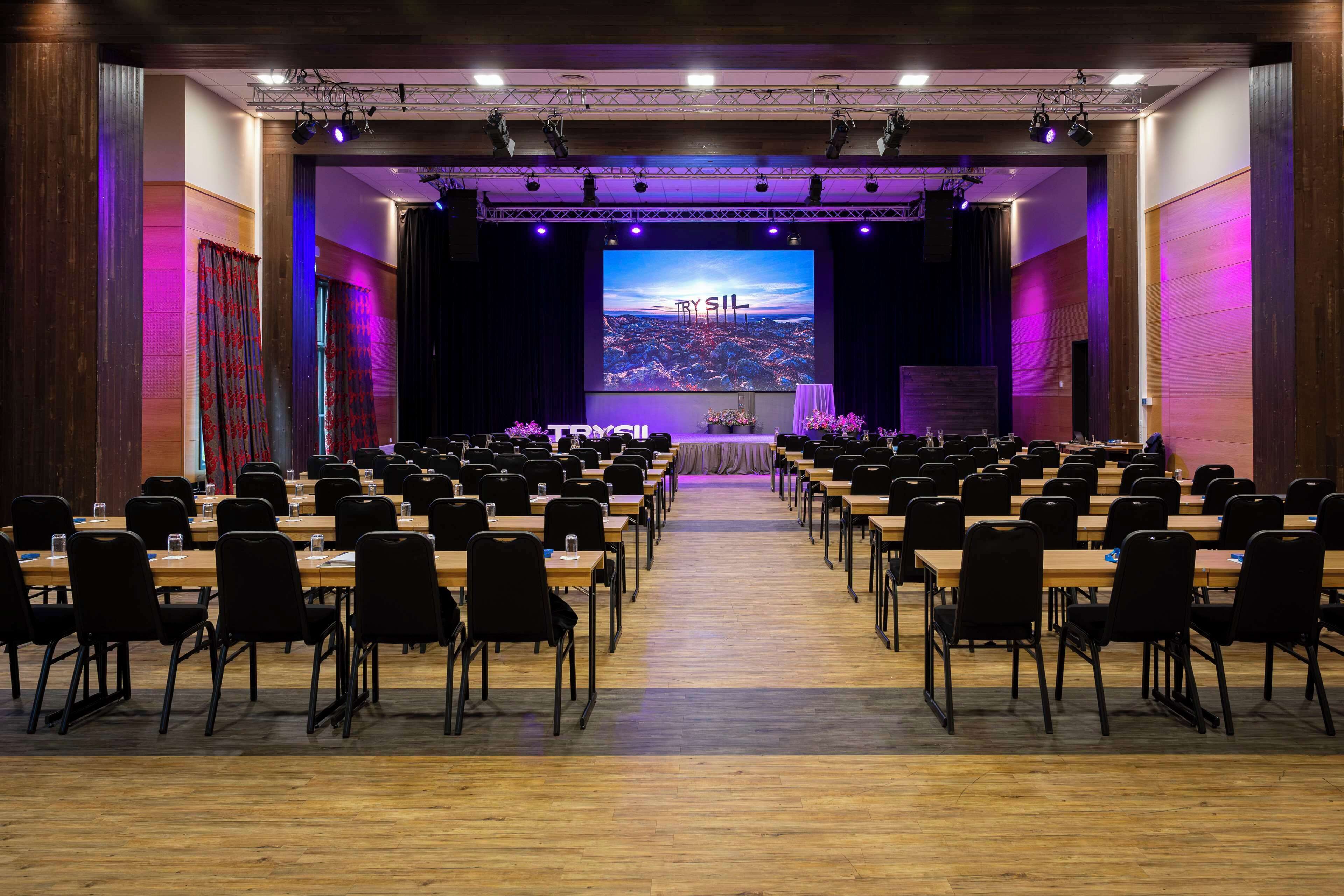 Un salón de conferencias con filas de mesas y sillas, un escenario con una pantalla grande que muestra 'TRY SIL', y un piso de madera.