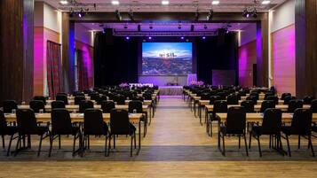 Un salón de conferencias con filas de mesas y sillas, un escenario con una pantalla grande que muestra 'TRY SIL', y un piso de madera.
