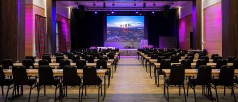 Un salón de conferencias con filas de mesas y sillas, un escenario con una pantalla grande que muestra 'TRY SIL', y un piso de madera.