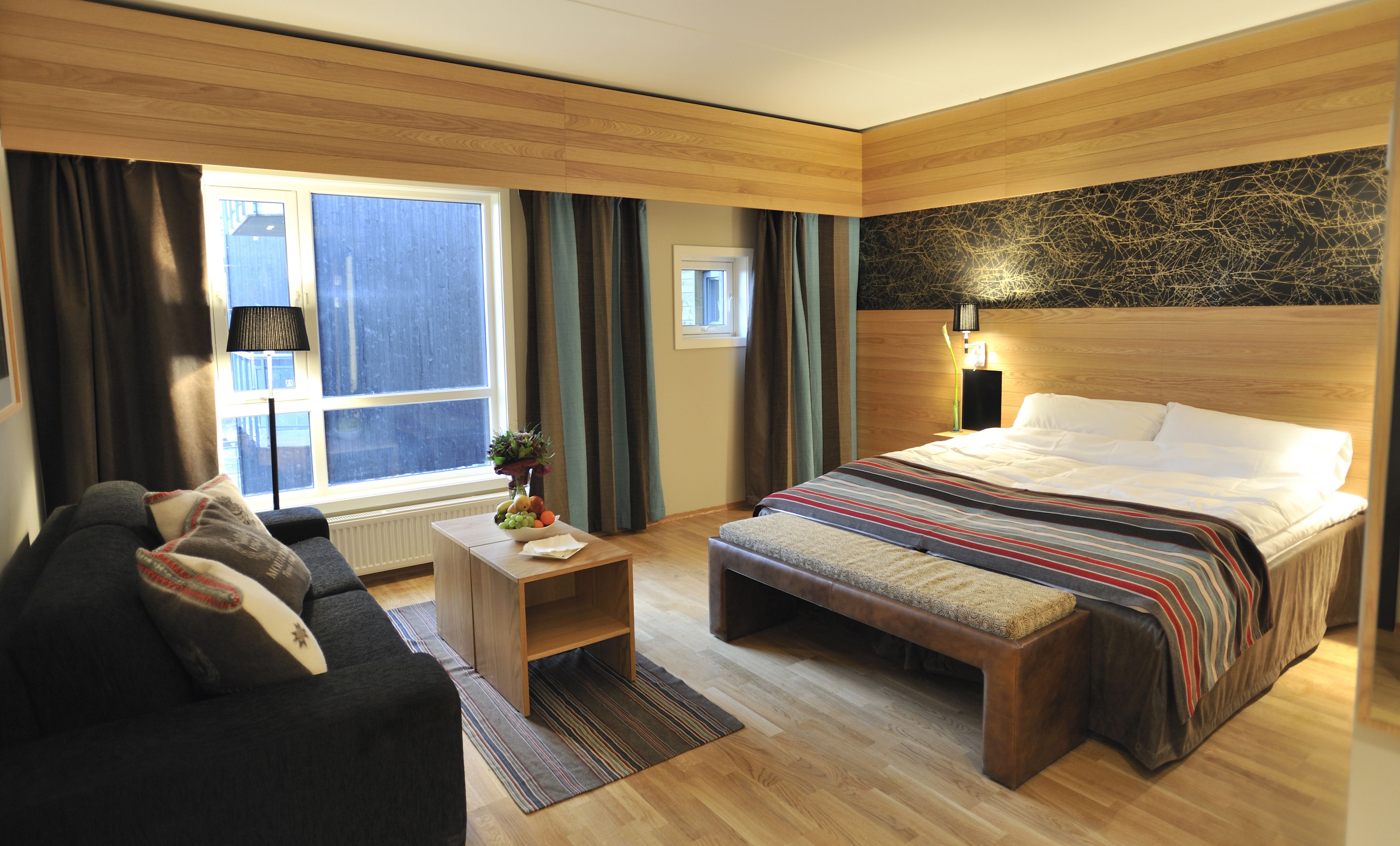 Una habitación de hotel moderna con suelo de madera, una cama grande con manta a rayas, un banco, una mesita con un jarrón de flores, un sofá y una ventana con cortinas.