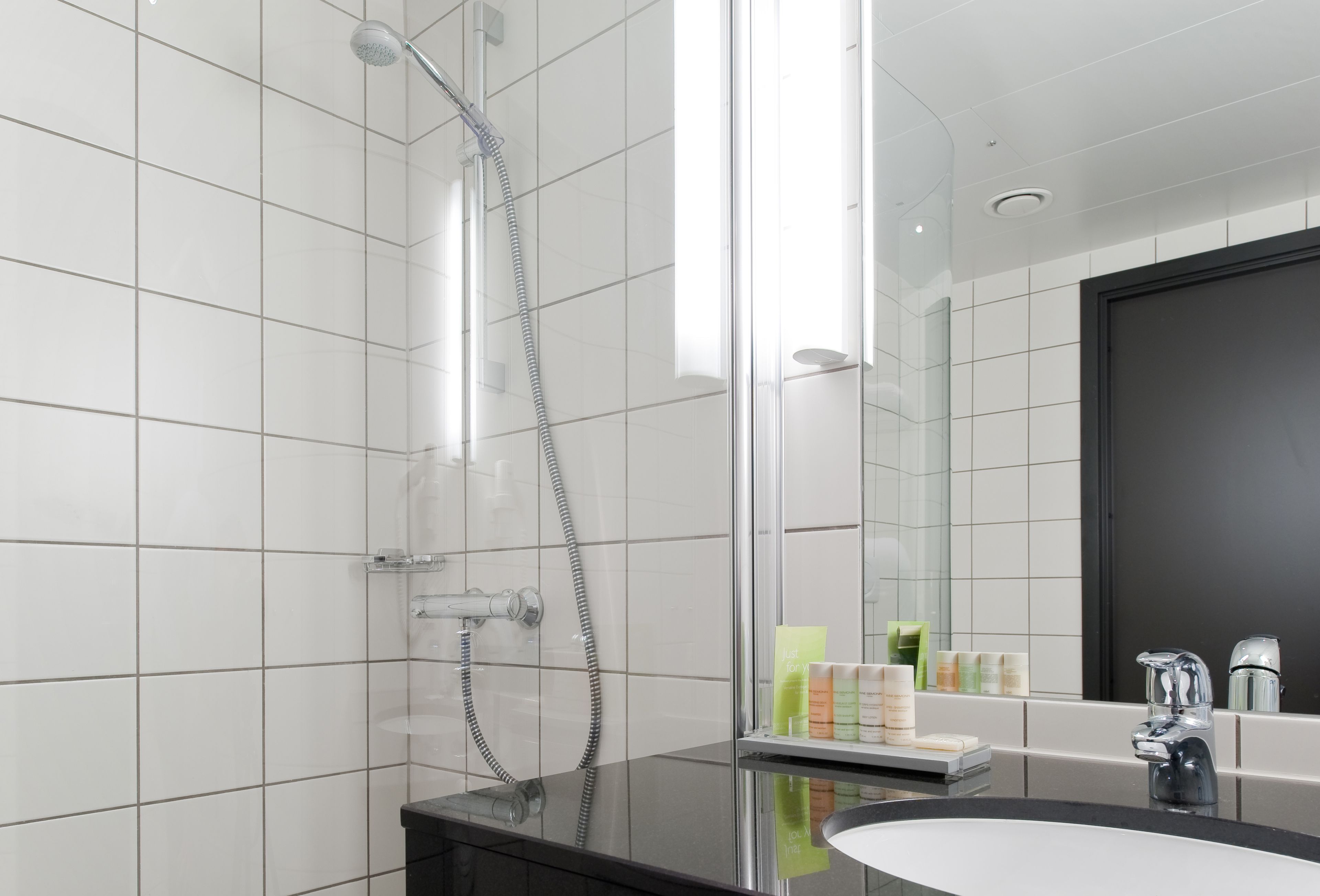 A modern bathroom with white tiled walls, a glass shower enclosure, a black vanity with a sink, and a mirror reflecting the bathroom.