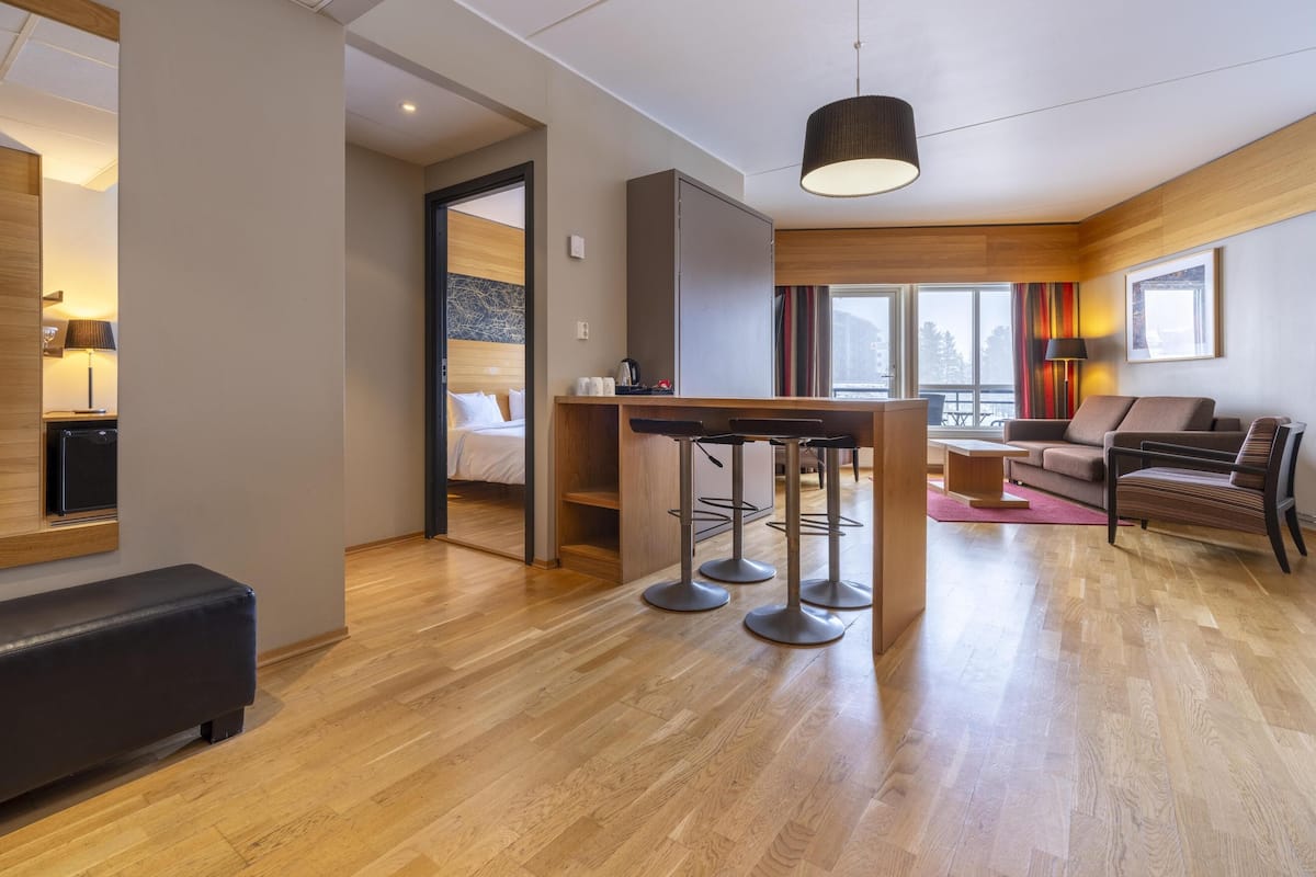 A modern hotel room with a wooden floor, a dining area with bar stools, a sofa, and a large window offering a view of the city.