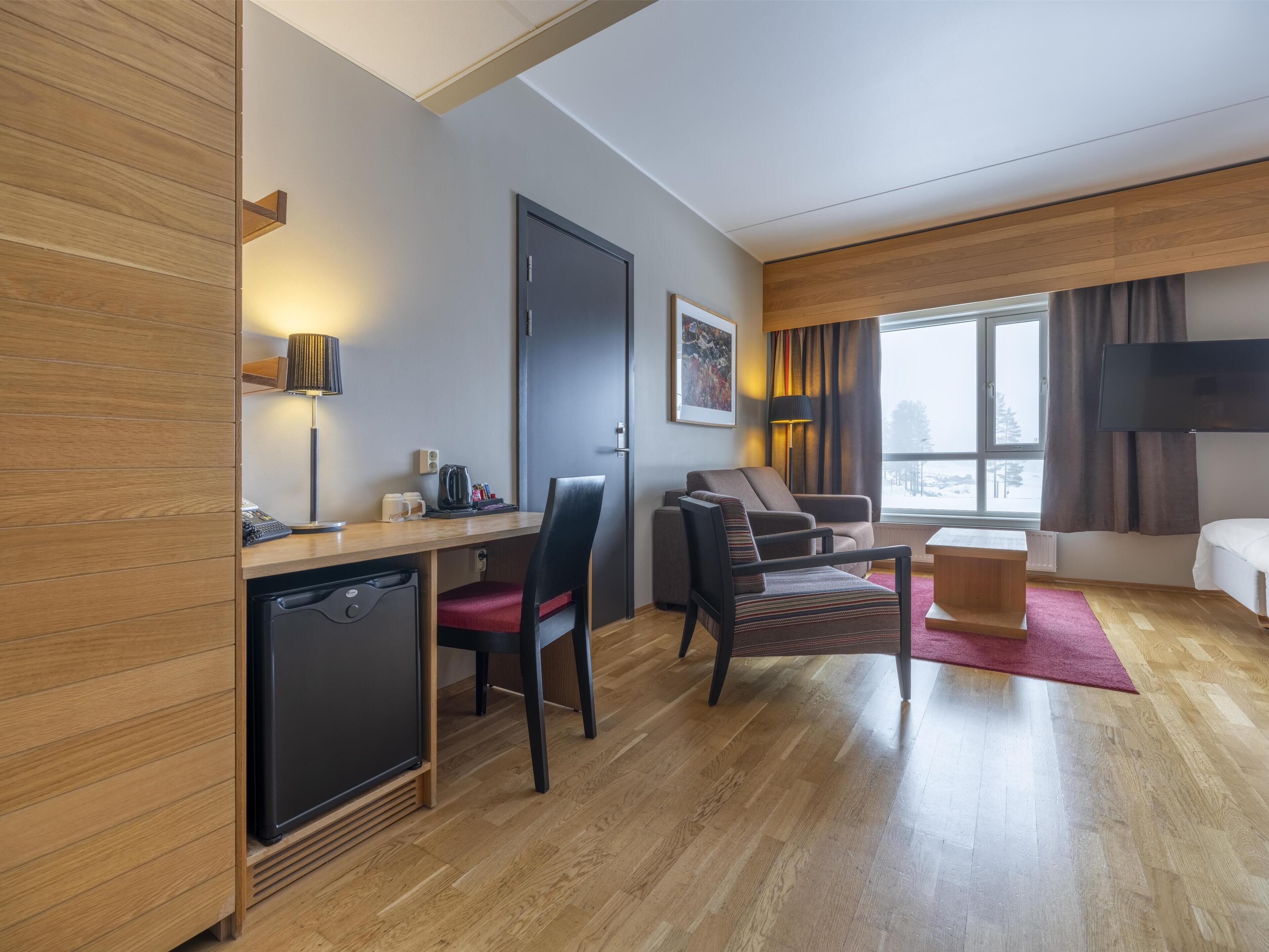 Quarto de hotel moderno com piso de madeira, uma mesa com cafeteira, um sofá, uma TV e uma janela com vista.