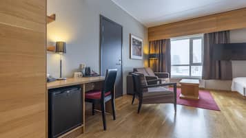 Quarto de hotel moderno com piso de madeira, uma mesa com cafeteira, um sofá, uma TV e uma janela com vista.
