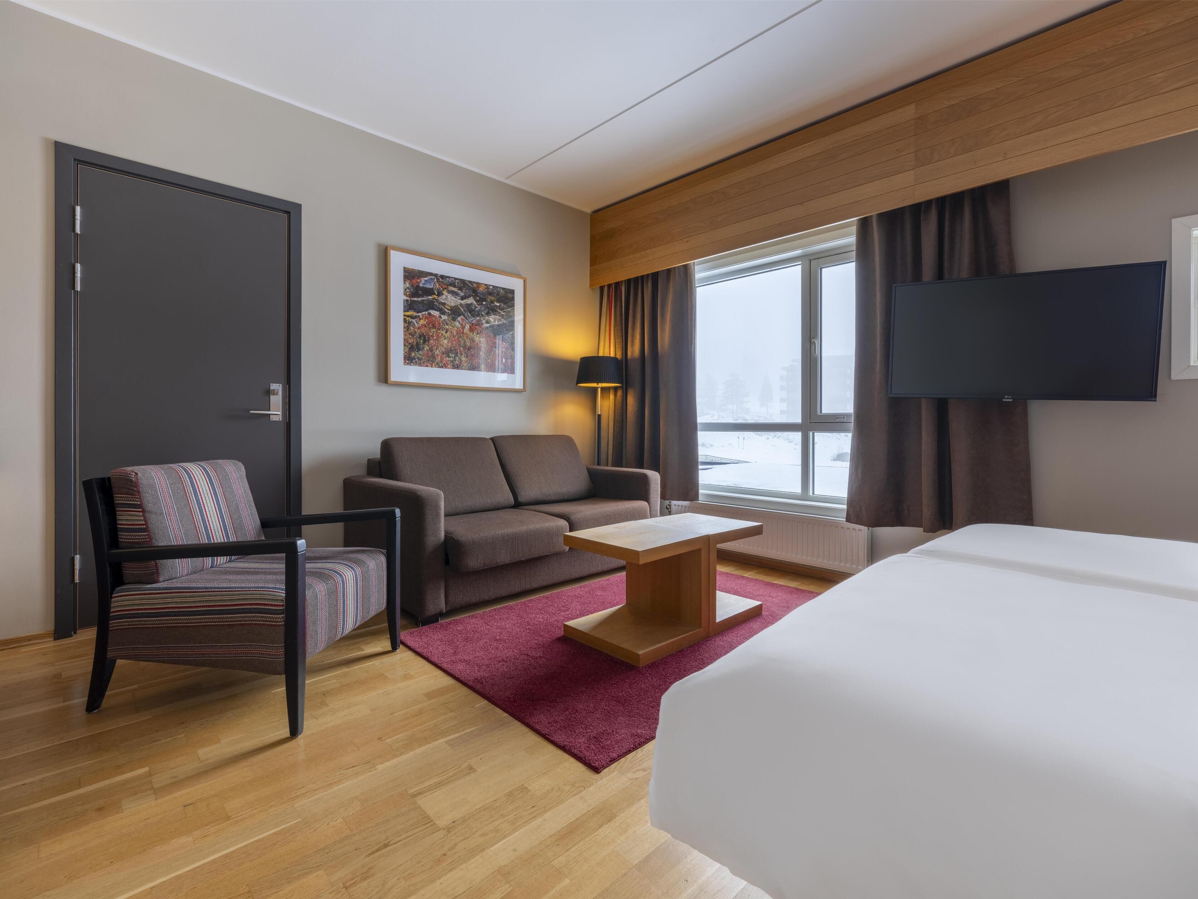 Ein Hotelzimmer mit Bett, einer Couch, einem Sessel, einem kleinen Tisch, einem Fernseher und einem Fenster mit Blick auf eine verschneite Landschaft.