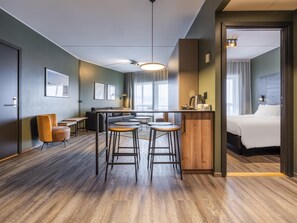 A modern hotel room with a wooden floor, a sofa, a dining area with stools, a kitchen area, and a large bed.