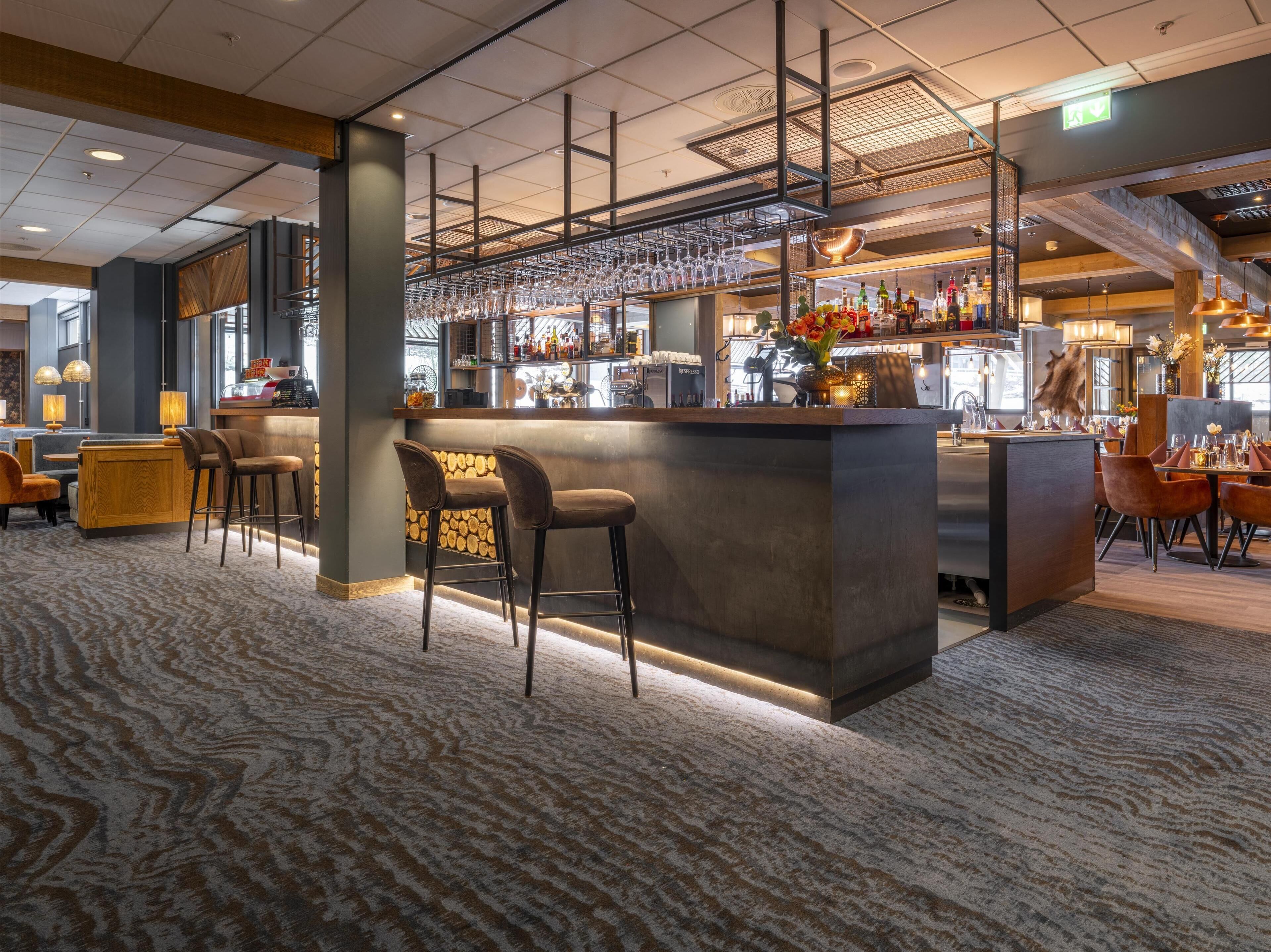 A bar area with stools, a counter with a coffee machine, and a variety of bottles and glasses.
