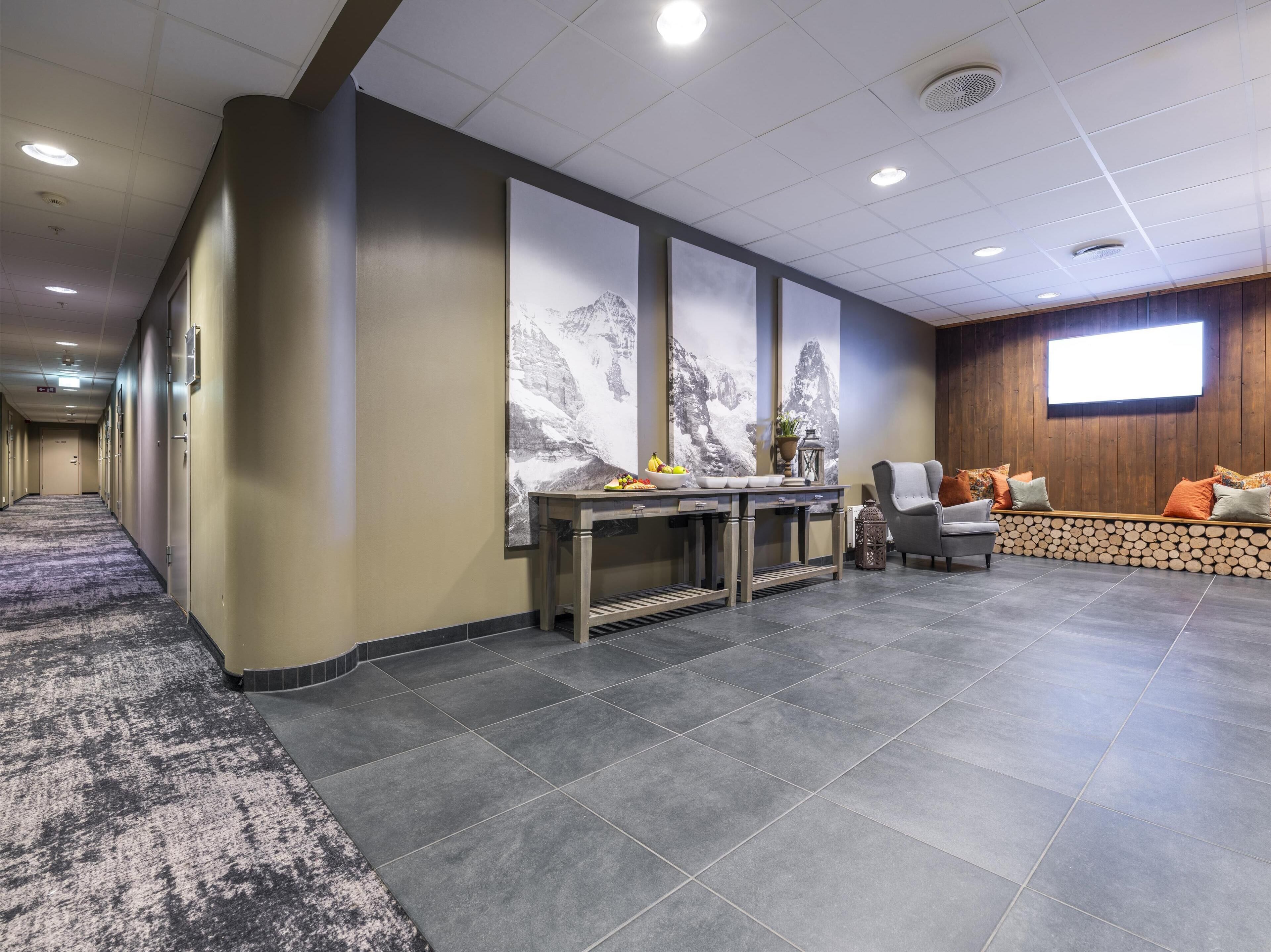 A hotel lobby with a long hallway, a reception desk with a vase and fruit bowl, a seating area with a sofa and cushions, and a large screen on the wall.