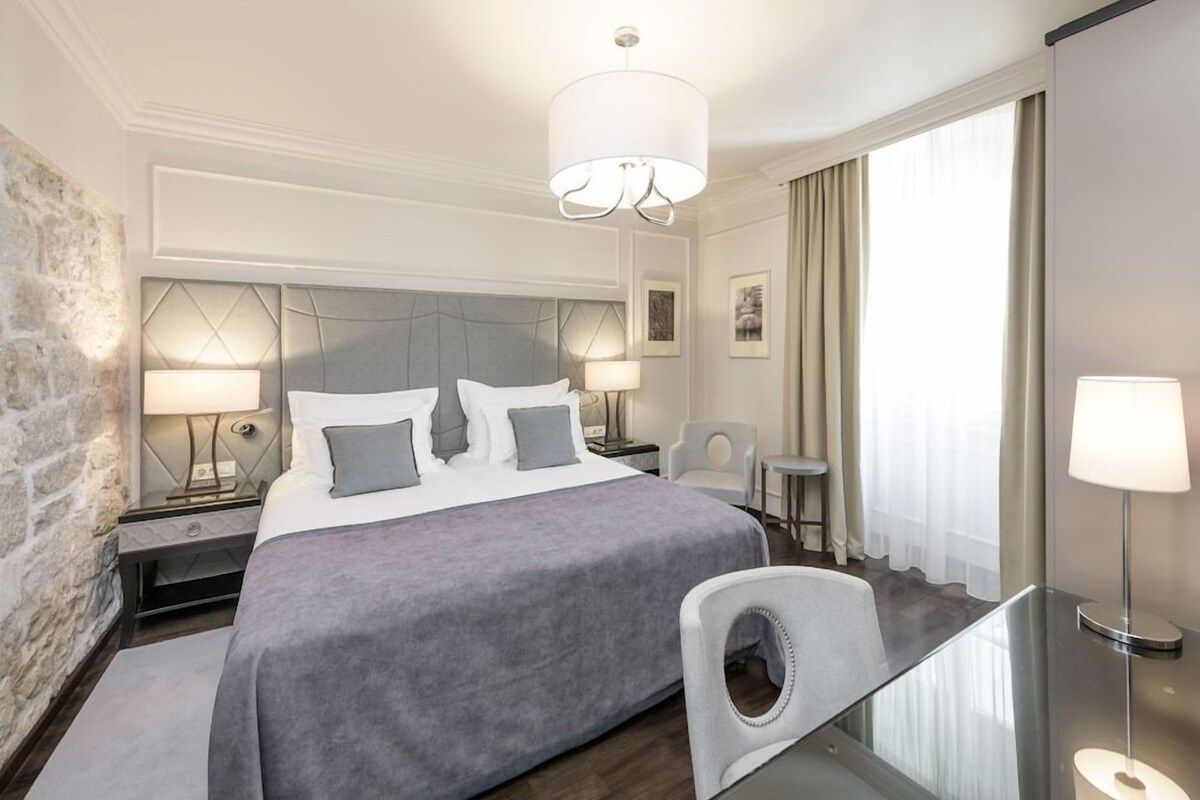 Een moderne hotelkamer met een groot bed, twee bedlampjes, een bureau met stoel, en een groot raam met gordijnen.