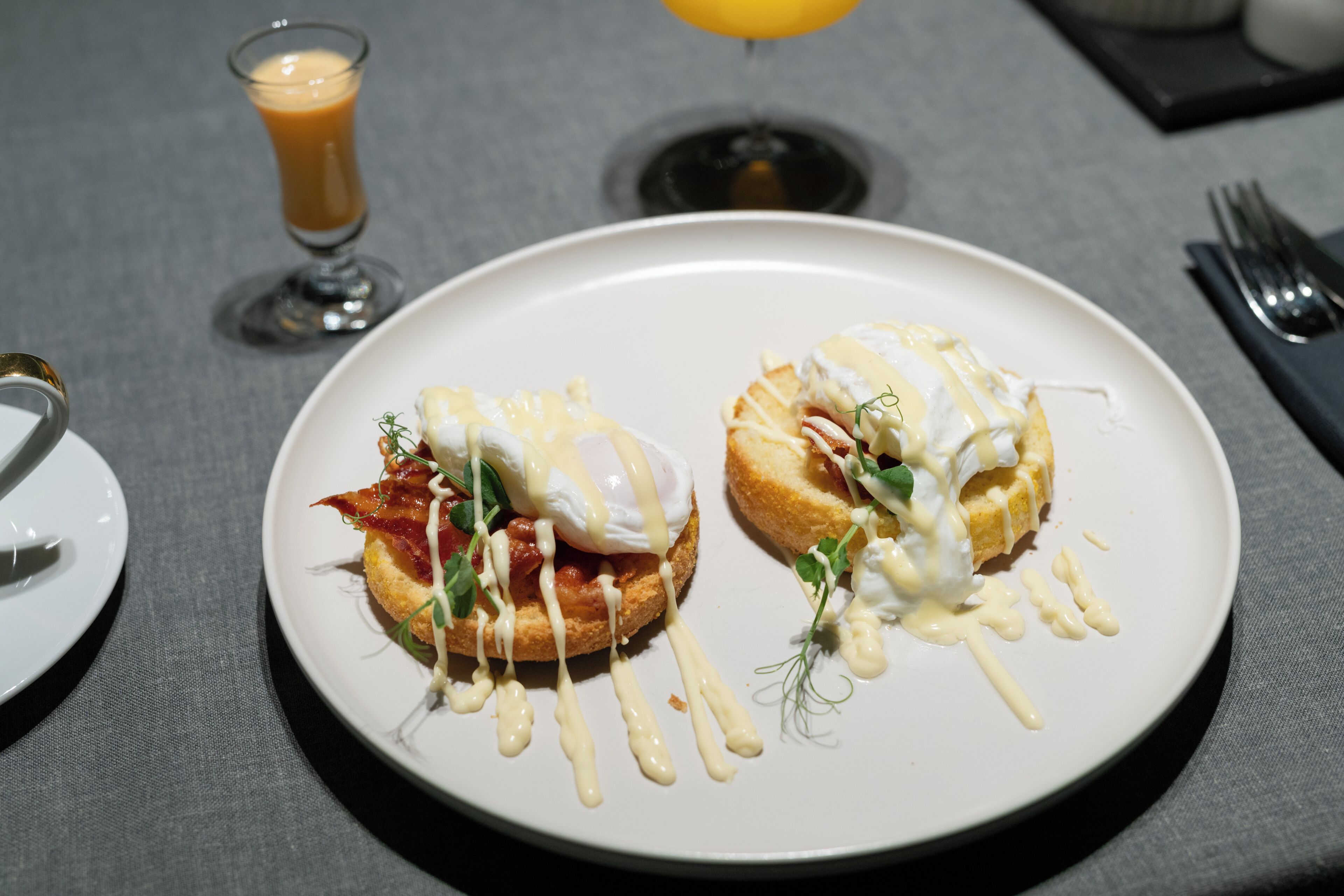 Un plato con dos sándwiches de huevos benedictinos y un vaso de jugo de naranja sobre una mesa.