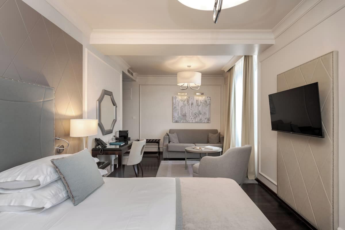 Ein Hotelzimmer mit einem großen Bett, einem Schreibtisch mit Stuhl, einer Sitzecke, einem Fernseher und einem Bild an der Wand.