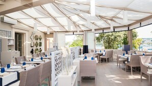 Un restaurante con terraza, mesas y sillas blancas, y vistas a la ciudad y al agua.