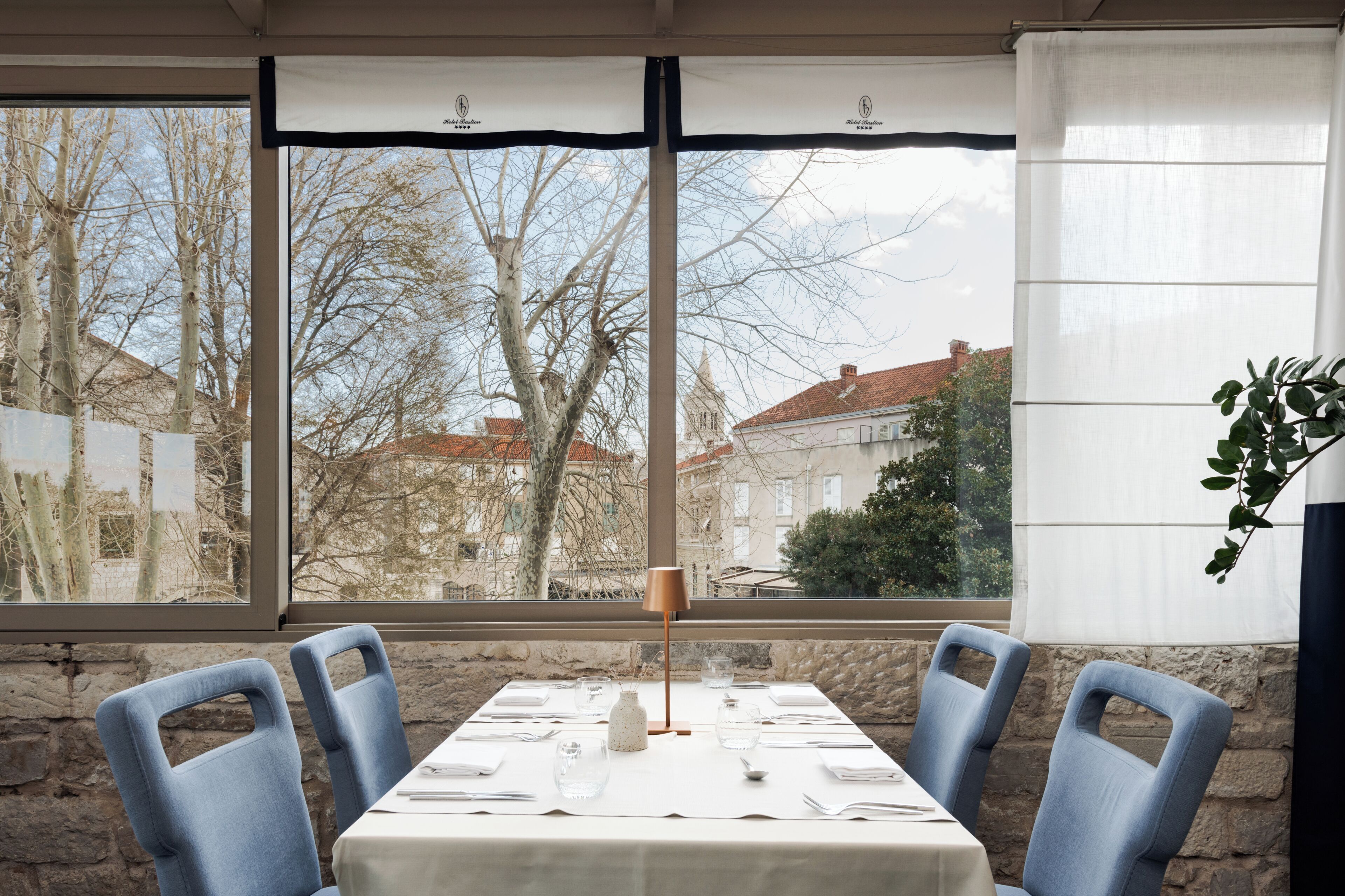 A dining table set with chairs, a lamp, and a view of buildings through large windows.