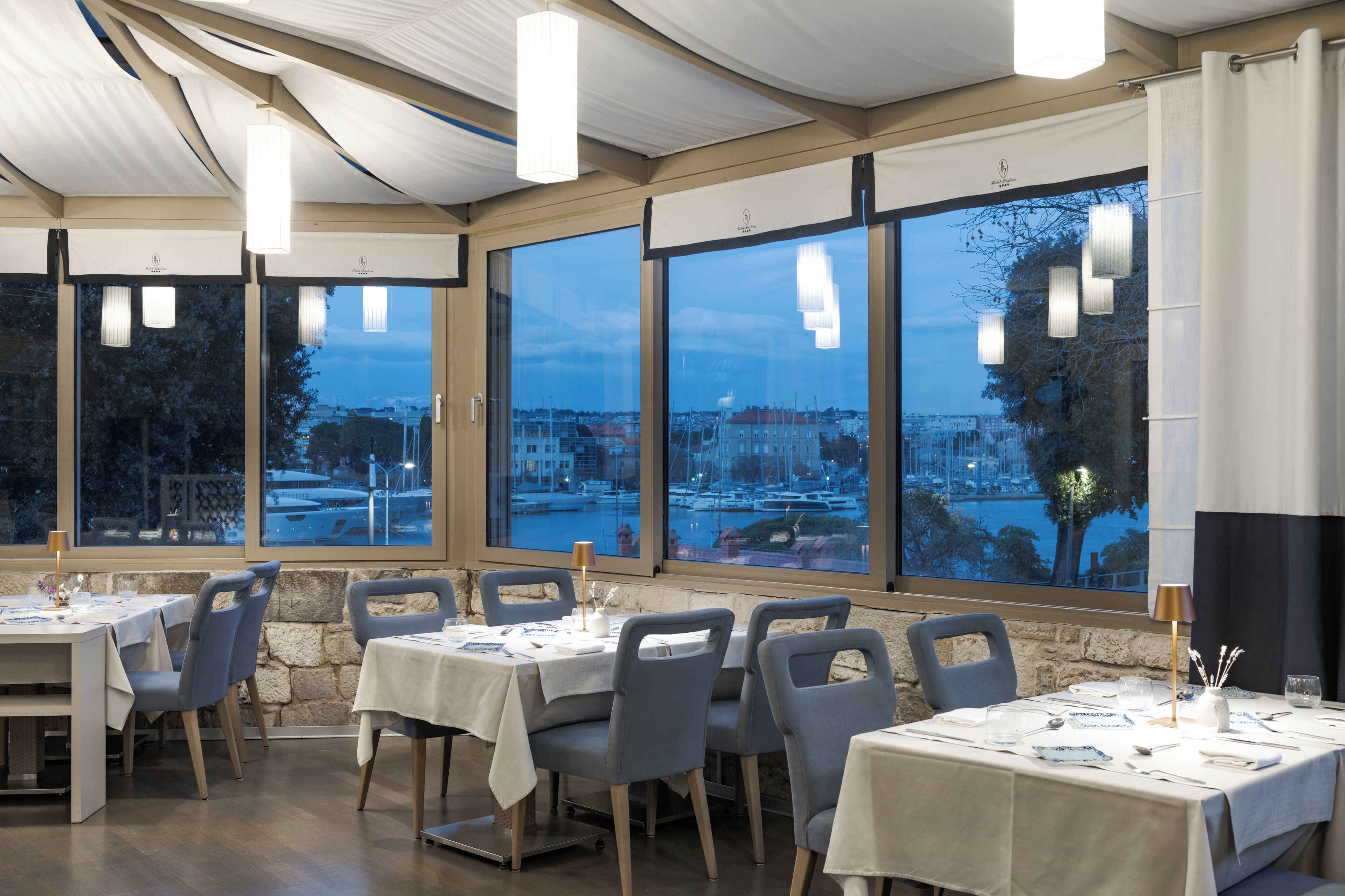 A restaurant with tables set for dining, large windows with a view of a marina, and pendant lighting.