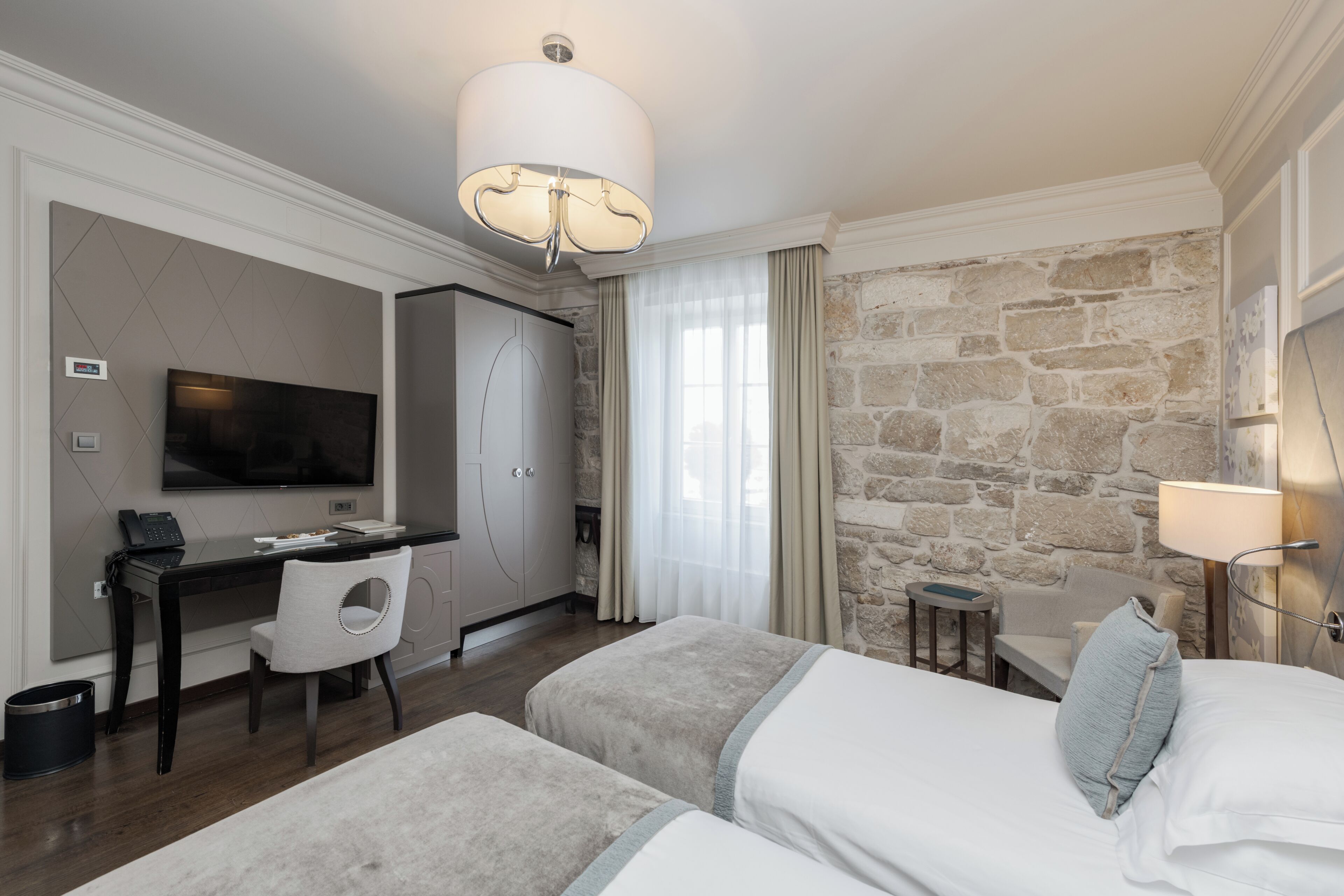 A hotel room with a bed, a desk, a chair, a television, and a stone wall.