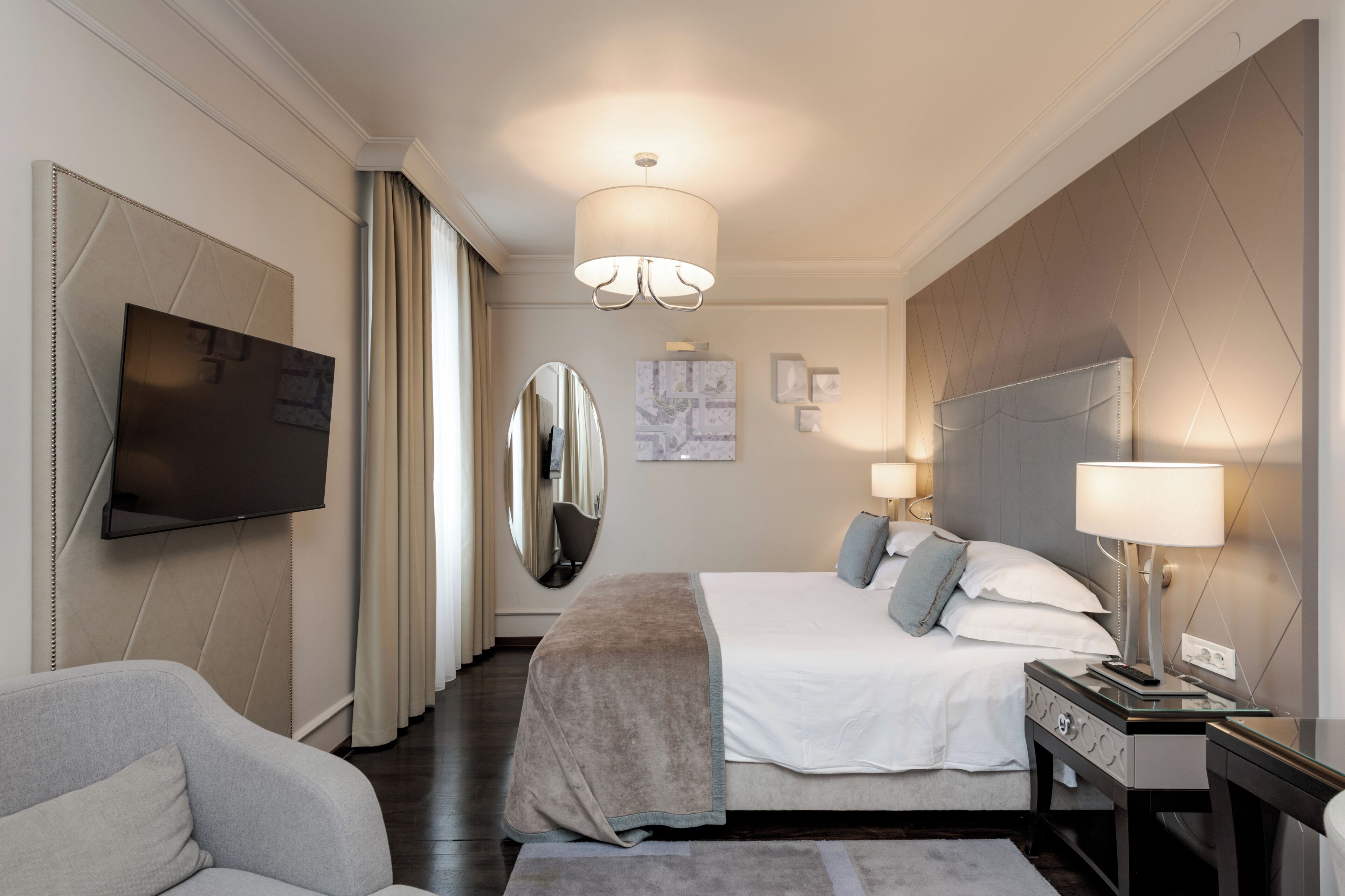 A modern hotel room with a large bed, a flat-screen TV, a mirror, and a desk with a lamp.